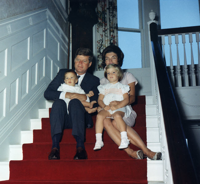 Photograph of President Kennedy and Jackie Kennedy sitting on the stairs with a red carpet runner. JFK Jr. sits in his father's lap, while Caroline Kennedy sits in her mother's lap.