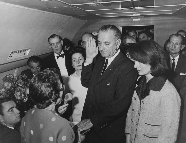 Black and white photograph of Lyndon Baines Johnson taking the presidential oath of office aboard air force one. Jackie Kennedy stands beside him