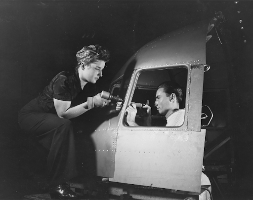 Black and white photograph of two riveters working on a plane at an aircraft company. They face each other while riveting a panel of the plane