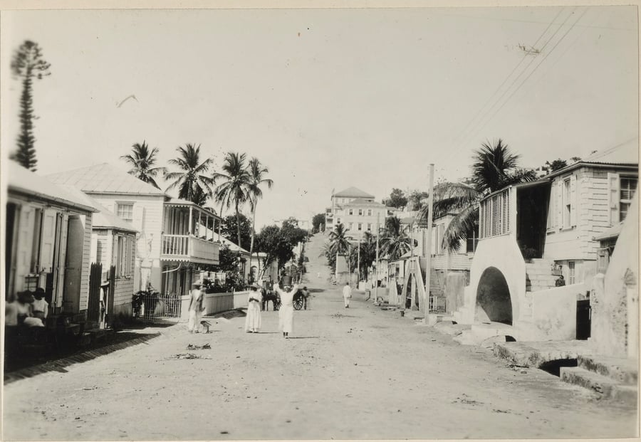 Black and white photograph of a street scene in St. Thomas, Virgin Islands. Several people walk down the street