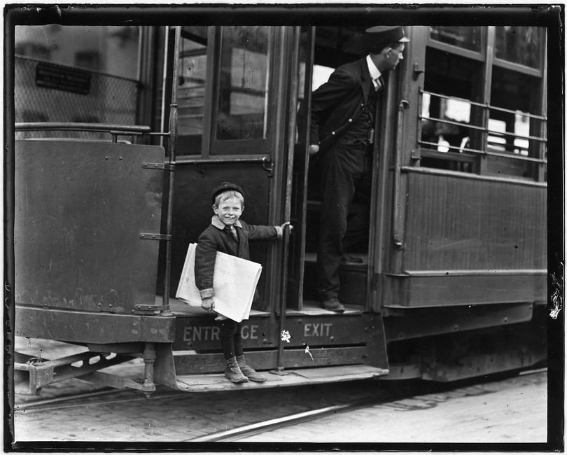 Black and white photograph of a young boy smiling at the camera and holding a stack of newspapers under his arm. He stands at the entrance of a street car, holding on with one hand while the conductor looks down the street.
