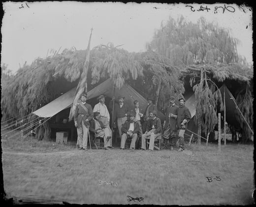 Black and white photograph of a camp scene. A tent covered in greens and leaves and branches is in the background, while a group of soldiers stand in front of it. 