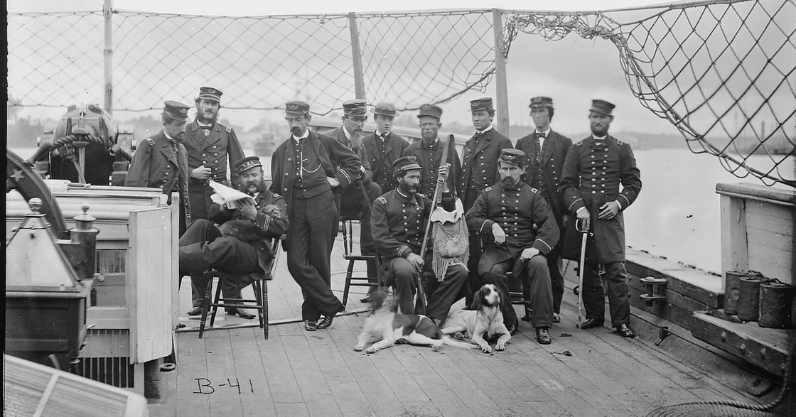 Black and white photograph of a group of men in uniform on the deck of a ship. Several men sit in chairs, and two dogs sit at their feet. 