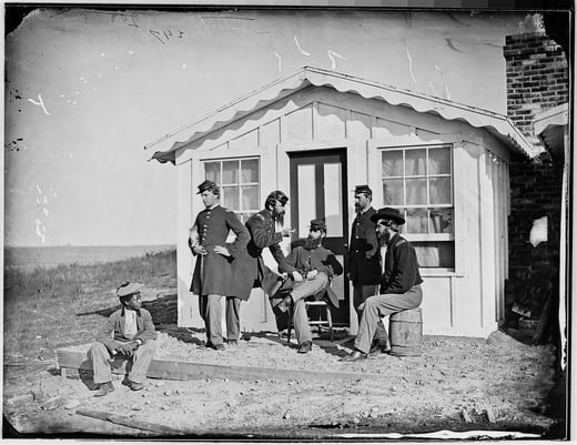 Black and white photograph of a group of soldiers sitting and standing in front of a small house.  A young boy sits off the the side.