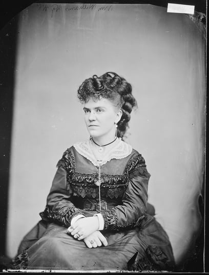 Black and white studio photograph of a woman posed in a dress with her hands crossed in her lap. 