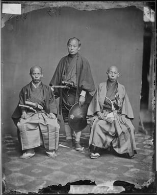 Black and white studio photograph of a group of three Japanese people. Two are seated, and one person in the middle stands. 