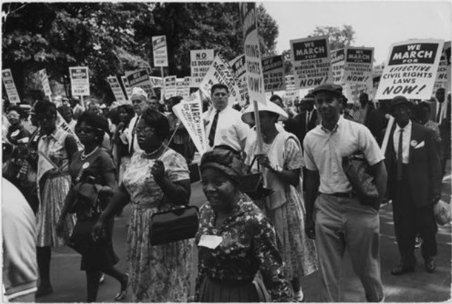 Black and white photograph of a group of people marching at the Civil Rights March on Washington. Some hold signs and smile at the camera