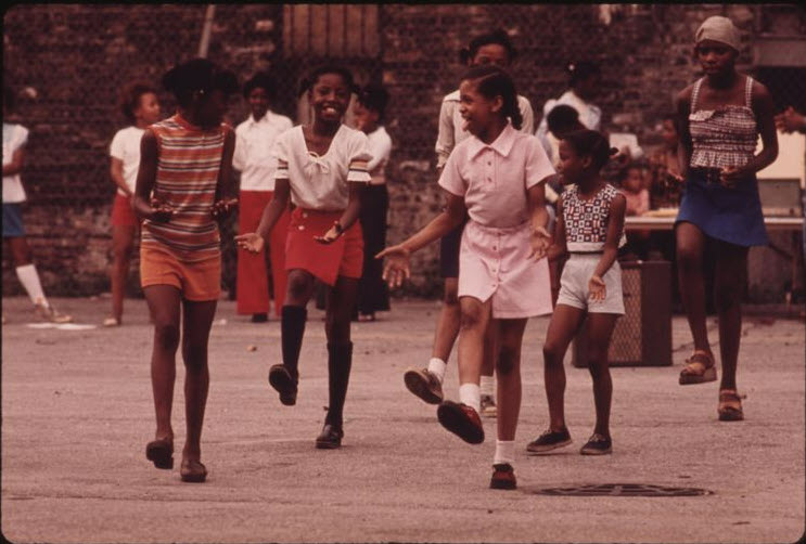 Group of young girls perform a dance routine outside, while smiling and laughing.