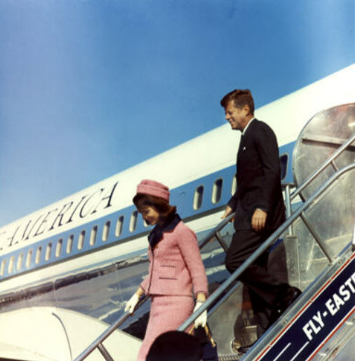 President and Mrs. Kennedy smile as they walk down the stairs from Air Force One. Mrs. Kennedy is wearing a pink suit and hat. Air Force One and a bright blue sky is behind them.
