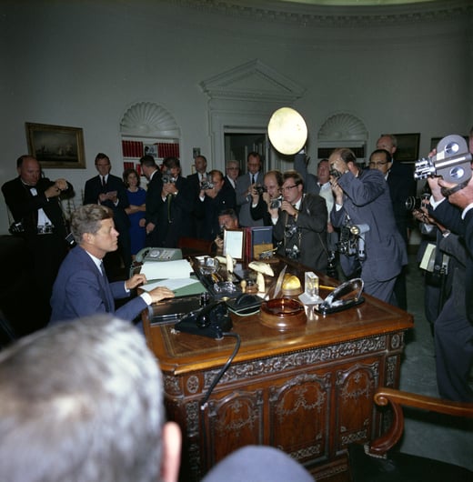 Photograph of President John F. Kennedy at his desk in the Oval Office about to sign the Cuba Quarantine Order. A crowd of reporters and photographers look on.