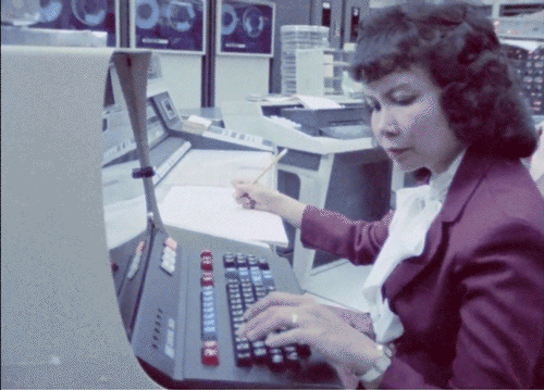 Animated gif of a woman in a red suit typing into a computer with her left hand, while holding a pencil in her right hand