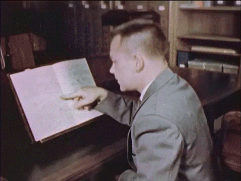 Animated GIF of a man in a suit reading a book on a stand. He points at the pages and then looks back to smile at someone out of the frame