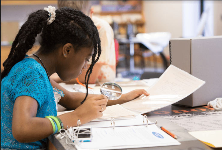 Side profile of a young girl sitting at a desk reviewing papers with a magnifying glasses in a 